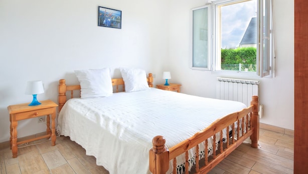 Un dormitorio con cama de madera, ropa de cama blanca, dos mesitas de noche con lámparas, una ventana con vistas a zonas verdes y un cuadro en la pared.