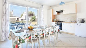 A dining area with a table set for four, a kitchen with white cabinets and a stove, and a view of the outside through large windows. - Les PerceNeige - Maison 85 m² à Port-Bail-Sur-Mer (Portbail)