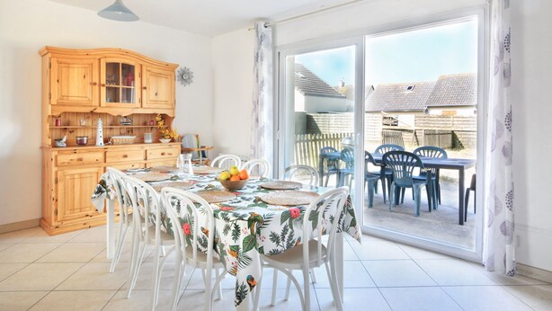 A dining area with a table set for four, surrounded by white chairs. A wooden cabinet with glass doors is to the left, and a sliding glass door leads to an outdoor patio with blue chairs. - Les PerceNeige - Maison 85 m² à Port-Bail-Sur-Mer (Portbail)
