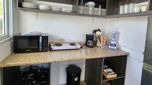 Cozinha compacta com micro-ondas, cafeteira e liquidificador na bancada. Prateleiras acima acomodam louças e recipientes.
