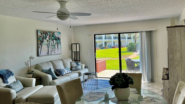 A spacious living room with a sofa, chairs, a coffee table, and a view of a well-maintained outdoor area with a lawn and patio furniture. - Modern St Augustine Beach Condo with Pool and Private Garage, Steps to Ocean. (St. Augustine)