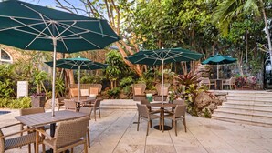 An outdoor patio area with wicker furniture and green umbrellas, surrounded by lush greenery and a stone staircase.