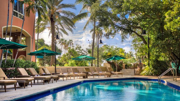 A swimming pool with lounge chairs and umbrellas, surrounded by palm trees and a building with a balcony. - Chic Condo near CocoWalk & Coral Gables (Miami)