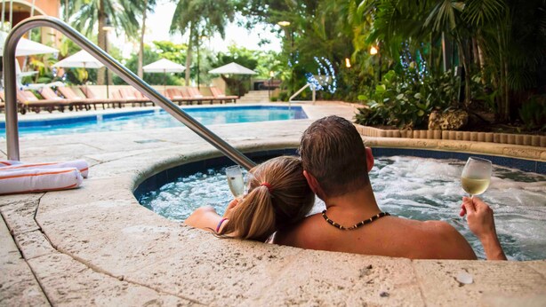 A couple relaxing in a hot tub with a pool in the background, surrounded by lounge chairs and umbrellas. - Bayfront Condo: Coconut Grove & Pool Access (Miami)