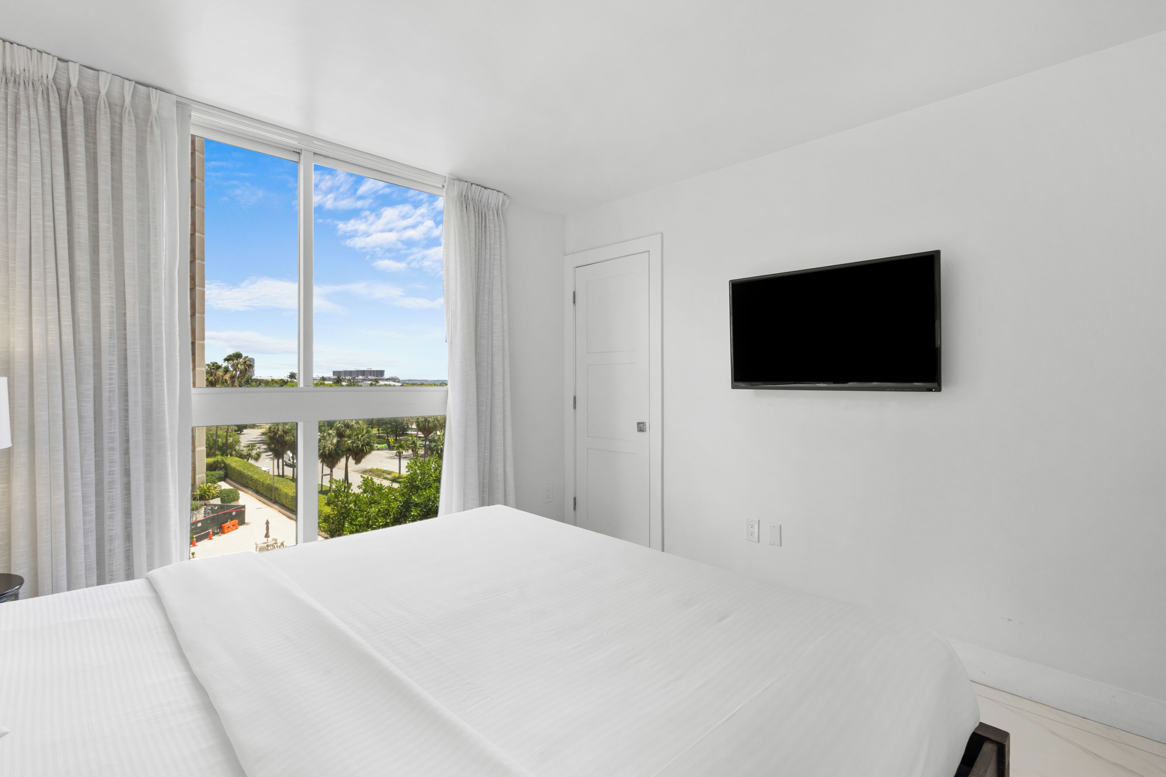 A hotel room with a large bed, a television on the wall, and a window with a view of a building and trees.