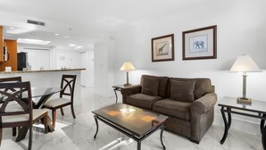 Una sala de estar moderna con una mesa de centro de vidrio, un sofá de cuero marrón y sillones a juego. Hay dos cuadros en la pared y dos lámparas sobre mesitas de noche.