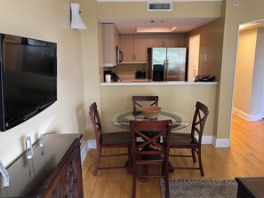 A dining area with a glass table, four wooden chairs, a television, and a kitchen in the background.
