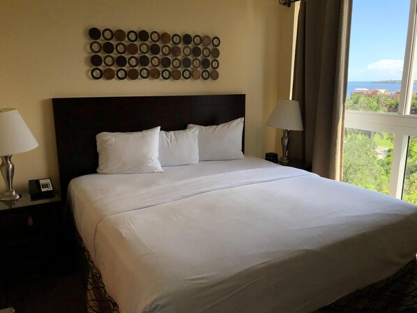 A hotel room with a large bed, two bedside lamps, a wall-mounted circular decoration, and a view of greenery and buildings through the window.