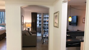 A modern hotel room with a living area, a sofa, a dining table, a kitchen with a dishwasher, and a balcony with a view of the city.