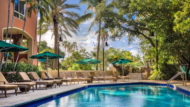 A swimming pool with lounge chairs and umbrellas, surrounded by palm trees and a building with a balcony.