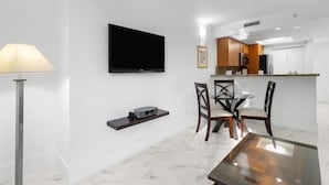 A modern living room with a flat-screen TV, a dining area with a glass table, and a kitchen with wooden cabinets.