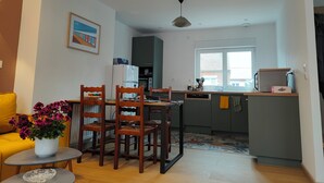 Un espace de vie compact comprenant un coin repas, une cuisine et un petit coin salon avec un canapé jaune et une table agrémentée d’un vase de fleurs.