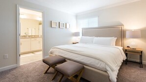 A bedroom with a large bed, a bedside table, a nightstand, a bench, and a door leading to a bathroom. - Last Minute Disney Getaway! Townhouse with Private Pool. Book Now! #118113 (Orlando)