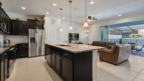 A modern kitchen with dark cabinets, stainless steel appliances, and a large island. There is a dining area with a brown sofa and a glass door leading to a balcony.