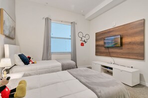 Ein Hotelzimmer mit zwei Betten, einem an einer Holzplatte befestigten Fernseher, einem Nachttisch mit Lampe sowie einer Wanddekoration mit Mickey Mouse.
