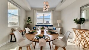 A dining area with a round table, white chairs, a blue sofa, and a view of the city through large windows. - Last Minute Disney Getaway! Luxury Condo with Private Balcony. Book Now! #65877 (Orlando)