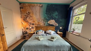 Une chambre avec un lit en bois, une fresque représentant une plage et un paysage sous-marin, une fenêtre décorée d’un motif en forme de cœur et une vue sur un phare.
