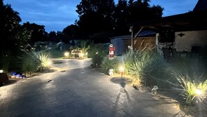 Un chemin pavé mène à un bâtiment sur lequel on peut voir une enseigne rouge et blanche, le tout étant entouré de verdure et éclairé par des projecteurs au sol.