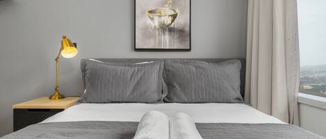 Un lit bien fait, avec une literie grise et deux serviettes blanches roulées, une lampe de bureau dorée et une œuvre d’art encadrée au-dessus du lit.