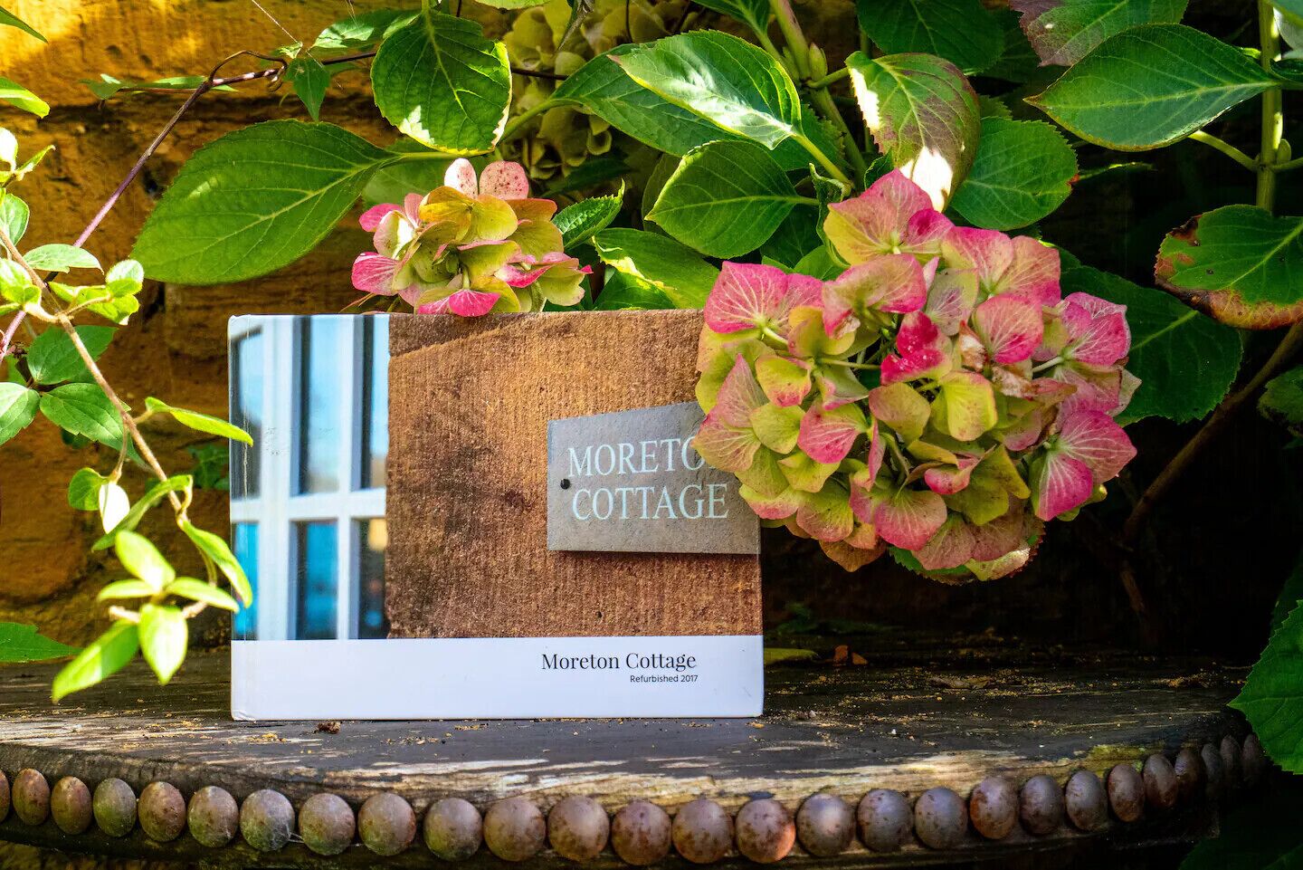 A sign for Moreton Cottage with a rustic wooden background and a window view.