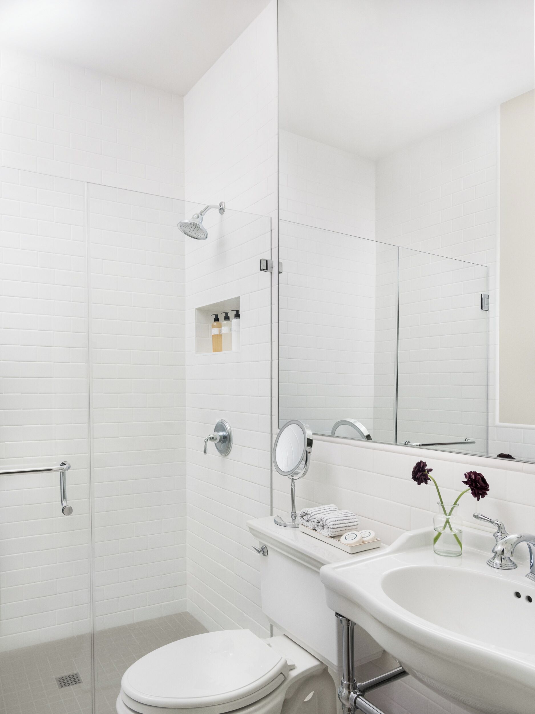 Un baño moderno con cabina de ducha de vidrio, un inodoro blanco, un lavabo con espejo y una jarra de vidrio con flores.
