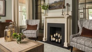 A cozy living room with a fireplace, two patterned armchairs, a coffee table with a vase, and a bookshelf.