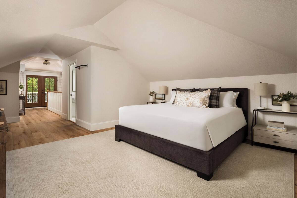 A spacious bedroom with a large bed, a nightstand, a lamp, and a view of a balcony.