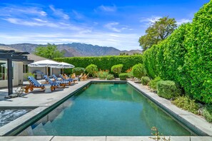 A rectangular swimming pool with clear water, surrounded by a low hedge and a row of blue and white striped lounge chairs.