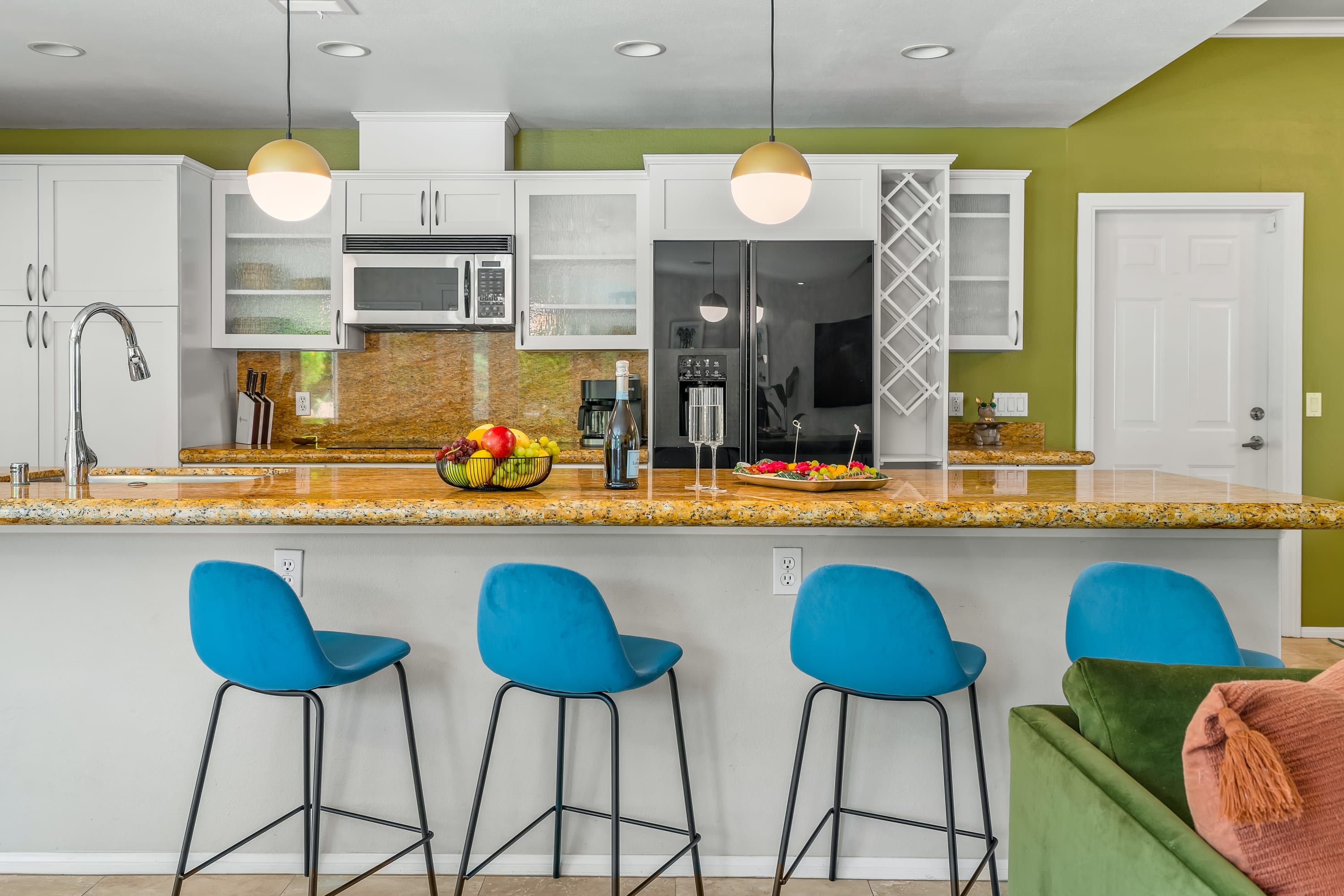 Una cocina moderna con armarios blancos, encimera de granito, microondas empotrado y refrigerador de acero inoxidable. Hay tres taburetes azules y un sofá verde en la zona de estar.