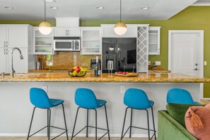 A modern kitchen with white cabinets, a granite countertop, a built-in microwave, and a stainless steel refrigerator. There are three blue bar stools and a green couch in the seating area.
