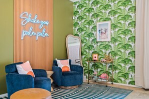 A room with two blue armchairs, a neon sign, a mirror, a small table with decorative items, and a patterned wallpaper.