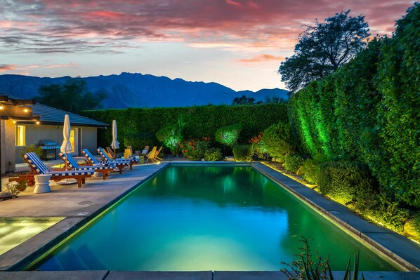 A rectangular pool with lounge chairs and umbrellas, surrounded by greenery and a mountain backdrop.