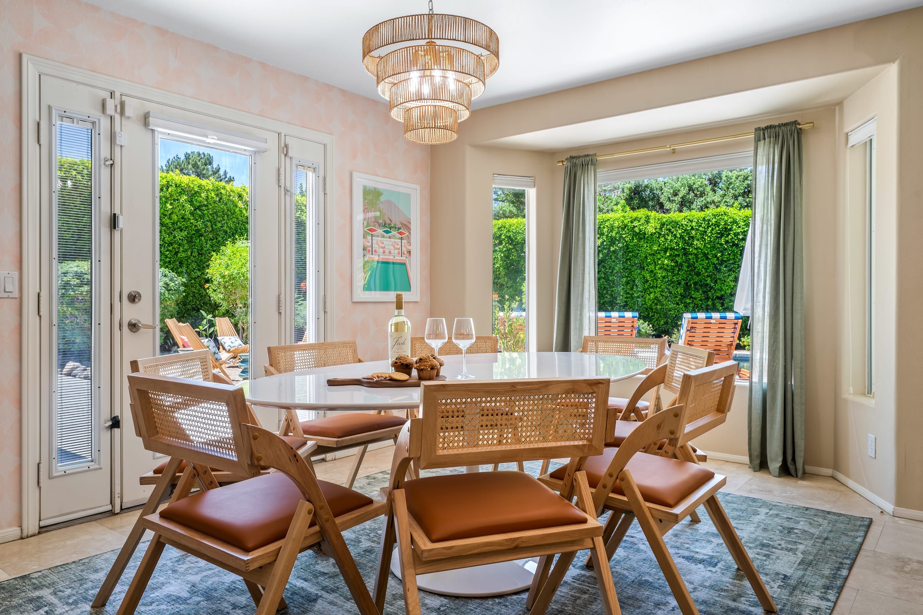 Un comedor con una mesa redonda, seis sillas de madera con cojines, una araña y vistas al exterior con vegetación.
