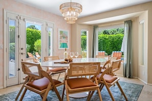 A dining area with a round table, six wooden chairs with cushions, a chandelier, and a view of greenery outside.