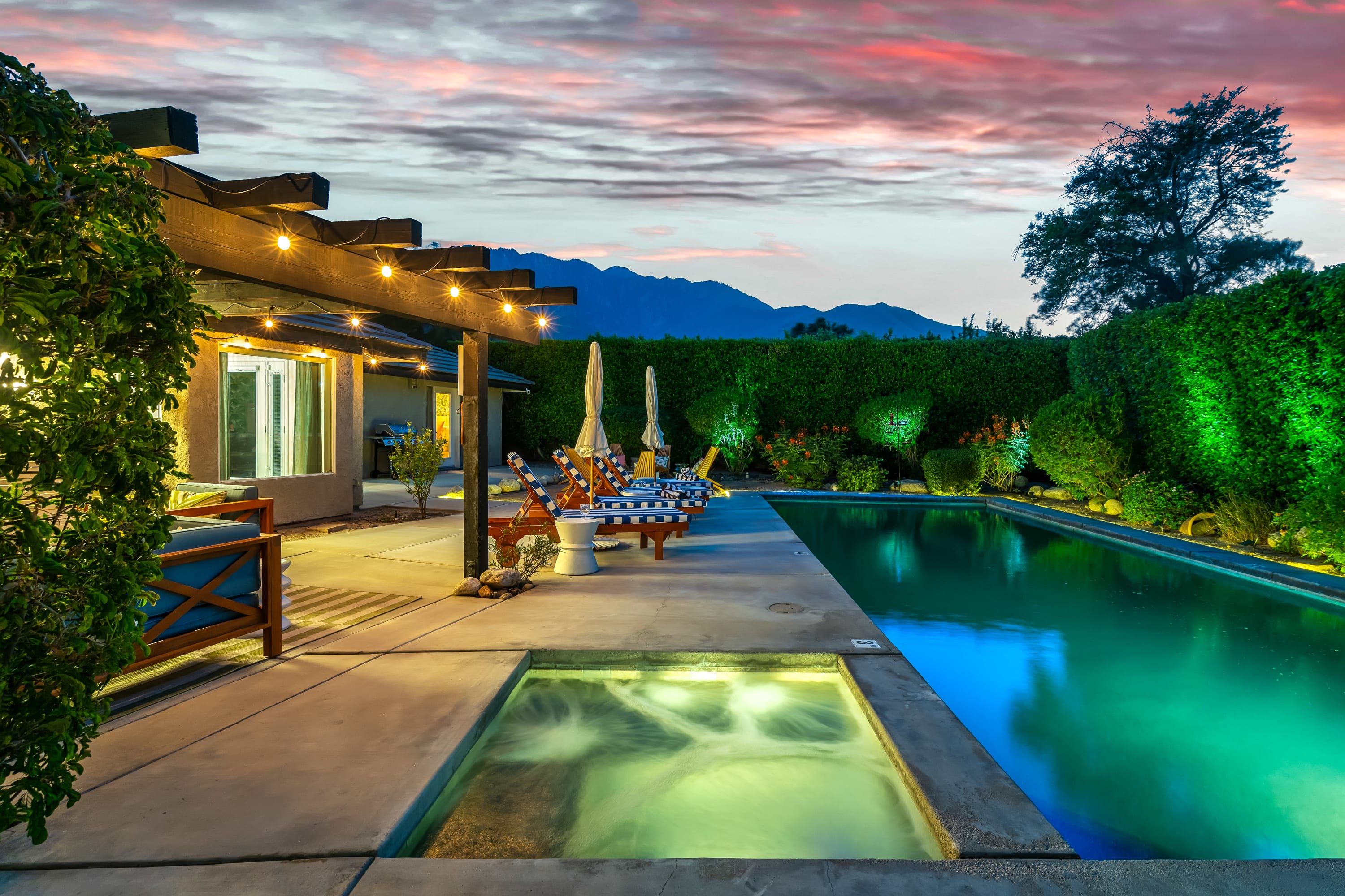 A pool area with lounge chairs and umbrellas, a pergola with string lights, and a view of mountains in the background.