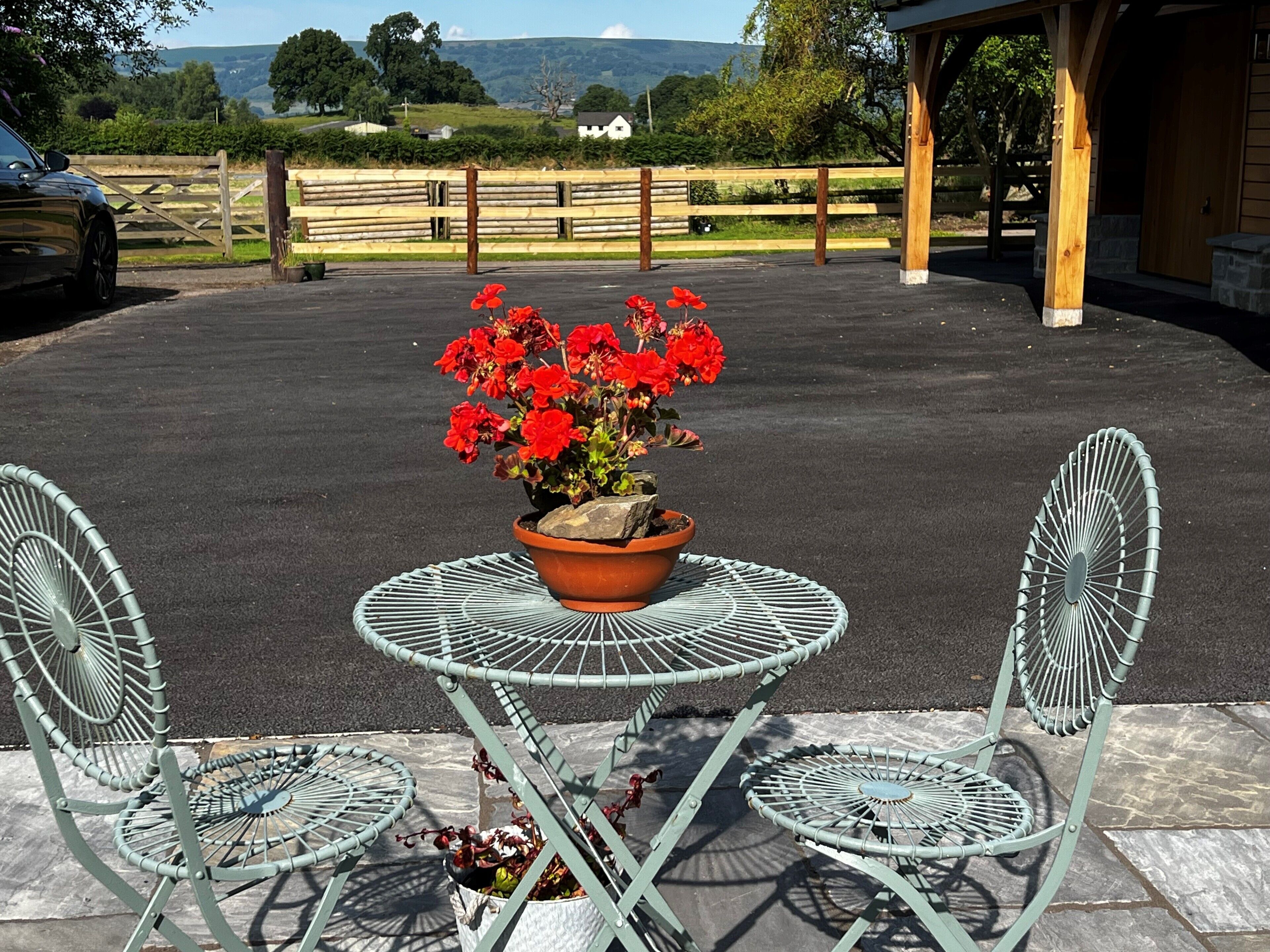 Un área de patio con una mesa redonda de metal y dos sillas, una planta en maceta con flores rojas y una valla de madera al fondo.