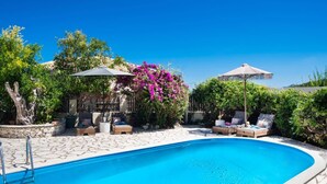 Une piscine entourée de verdure et dotée d’une terrasse en pierre, avec des chaises longues et des parasols.