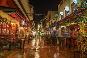 Une rue étroite bordée de boutiques et de cafés, avec un pavé mouillé et des guirlandes lumineuses suspendues.
