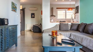 Ein Wohnzimmer mit einer Couch, einem Couchtisch und einem Fernseher. Im Hintergrund ist eine Küche mit einem Fenster und einer Hängelampe zu sehen.