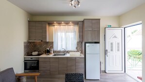A compact kitchen with wooden cabinets, a built-in oven, a refrigerator, and a window with curtains. - Estia Coastal Dream - Lush Enchanting Garden Gem (Pylos-Nestoras)