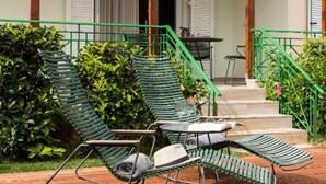 Una terraza de madera con dos sillones de jardín verdes y una mesa, un porche cubierto y una escalera que lleva a un edificio con contraventanas blancas.