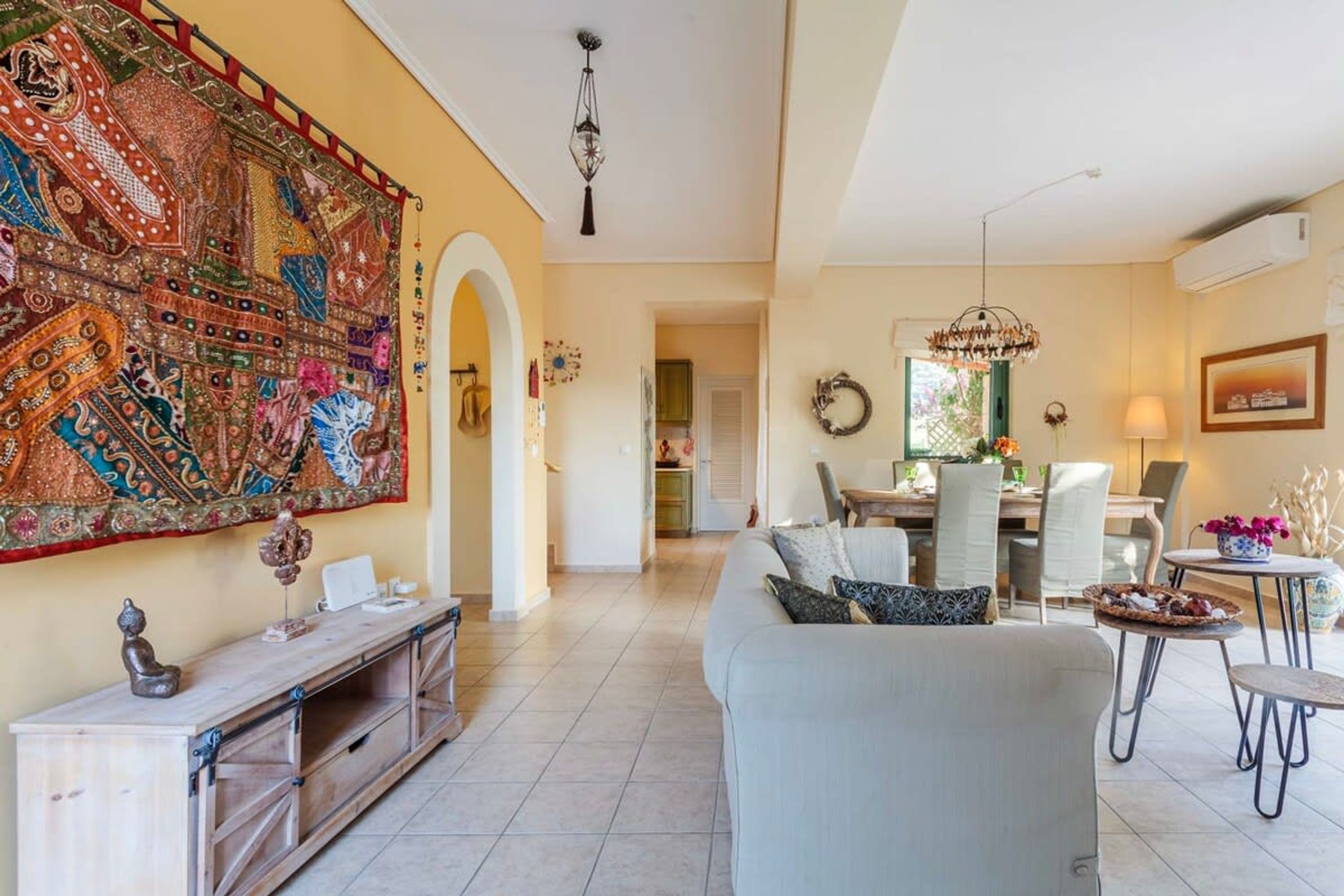 A spacious living room with a large sofa, a dining area with a chandelier, and a vibrant tapestry on the wall.