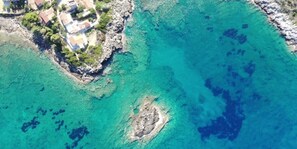 Vista aerea di una zona costiera con acque cristalline color turchese, coste rocciose e un gruppo di edifici con tetti rossi.