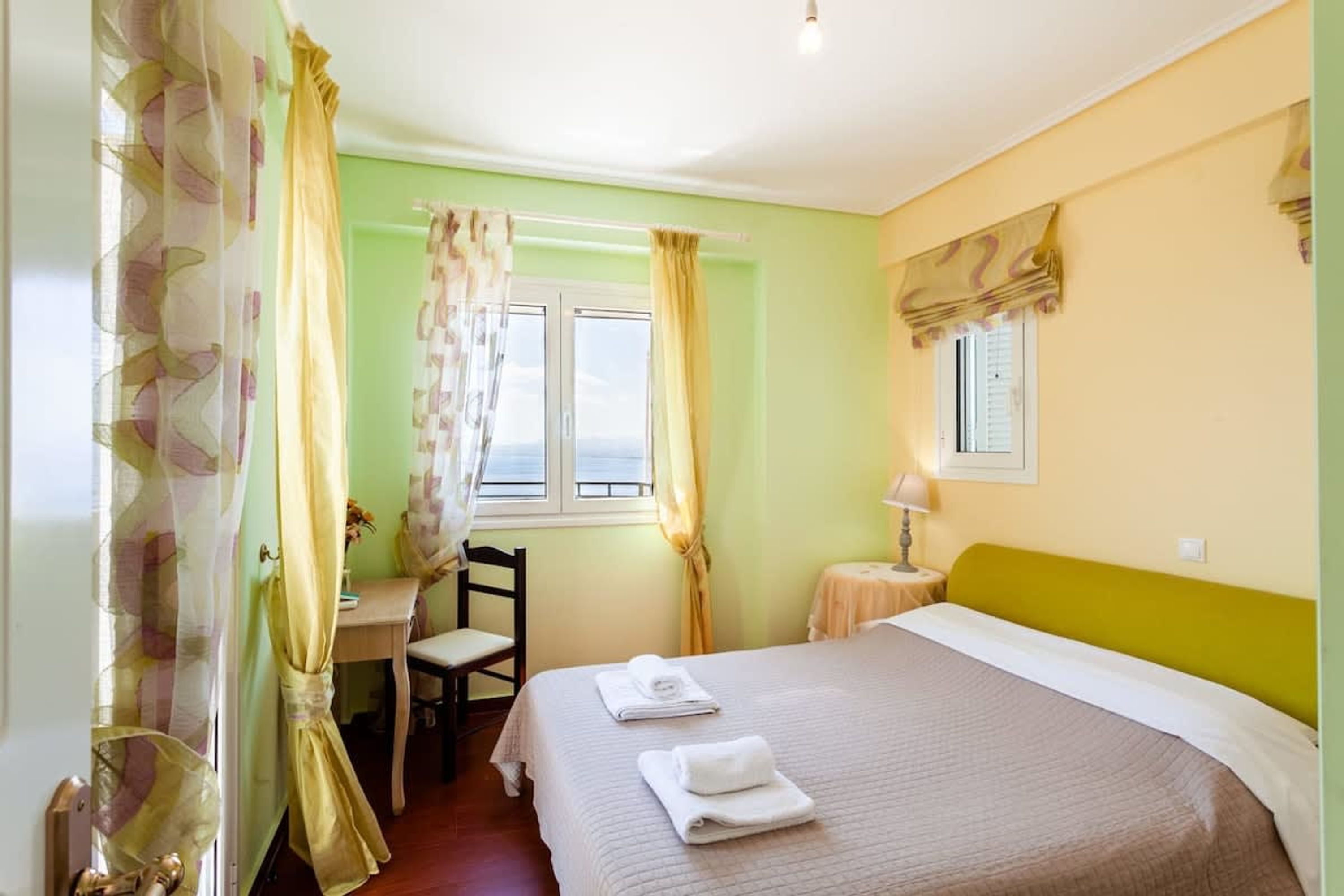 Quarto de hotel com cama, escrivaninha e cadeira. Há uma janela com cortinas, um abajur numa mesinha de cabeceira e vista para o exterior.