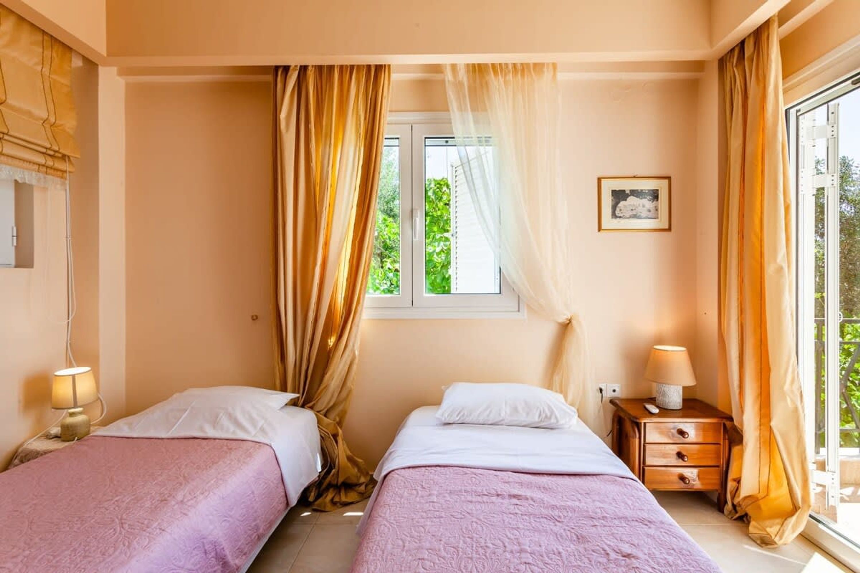 A bedroom with two beds, a window with curtains, a bedside table with a lamp, and a framed picture on the wall.