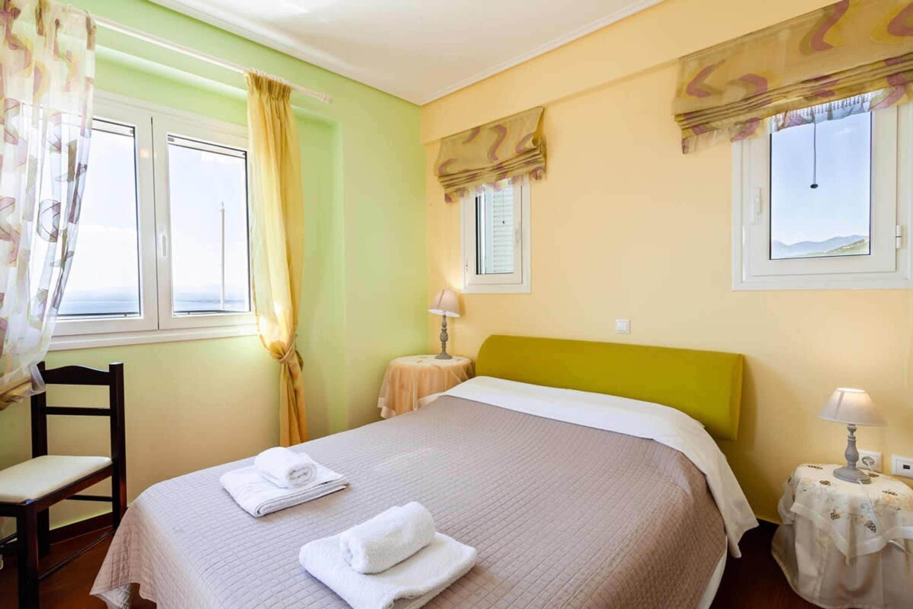 Een hotelkamer met een bed, een stoel, een nachtkastje met lamp en een raam met gordijnen.