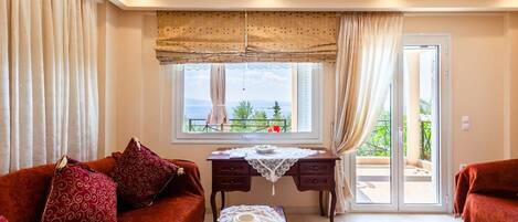 A cozy living room with a sofa, a coffee table, and a desk. There is a window with a view of greenery and a balcony.