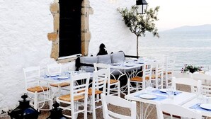 An outdoor dining area with white tables and chairs, set against a white wall with a view of the sea. - Thalassa Return To Timeless Spetses (Spetses)