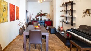 A dining area with a long wooden table, chairs, and a piano. There are artworks on the walls and a shelf with decorative items.