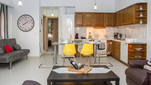 A modern living room with a sofa, a dining area with yellow chairs, a kitchen with wooden cabinets, and a wall clock. - Nina s Villa Cosy Seaview Retreat (Kalamata)